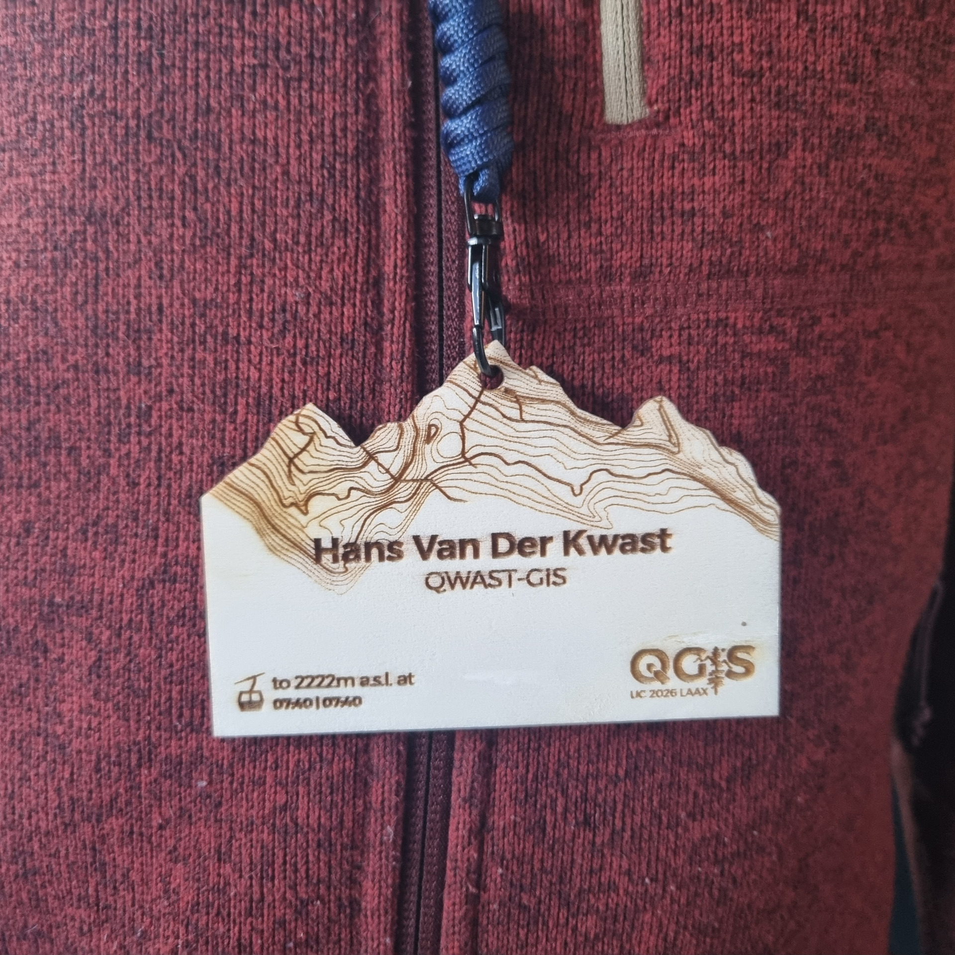 Close-up of a maroon fabric zipper with a creative name tag in the shape of mountains reading “Hans Van Der Kwast” attached.