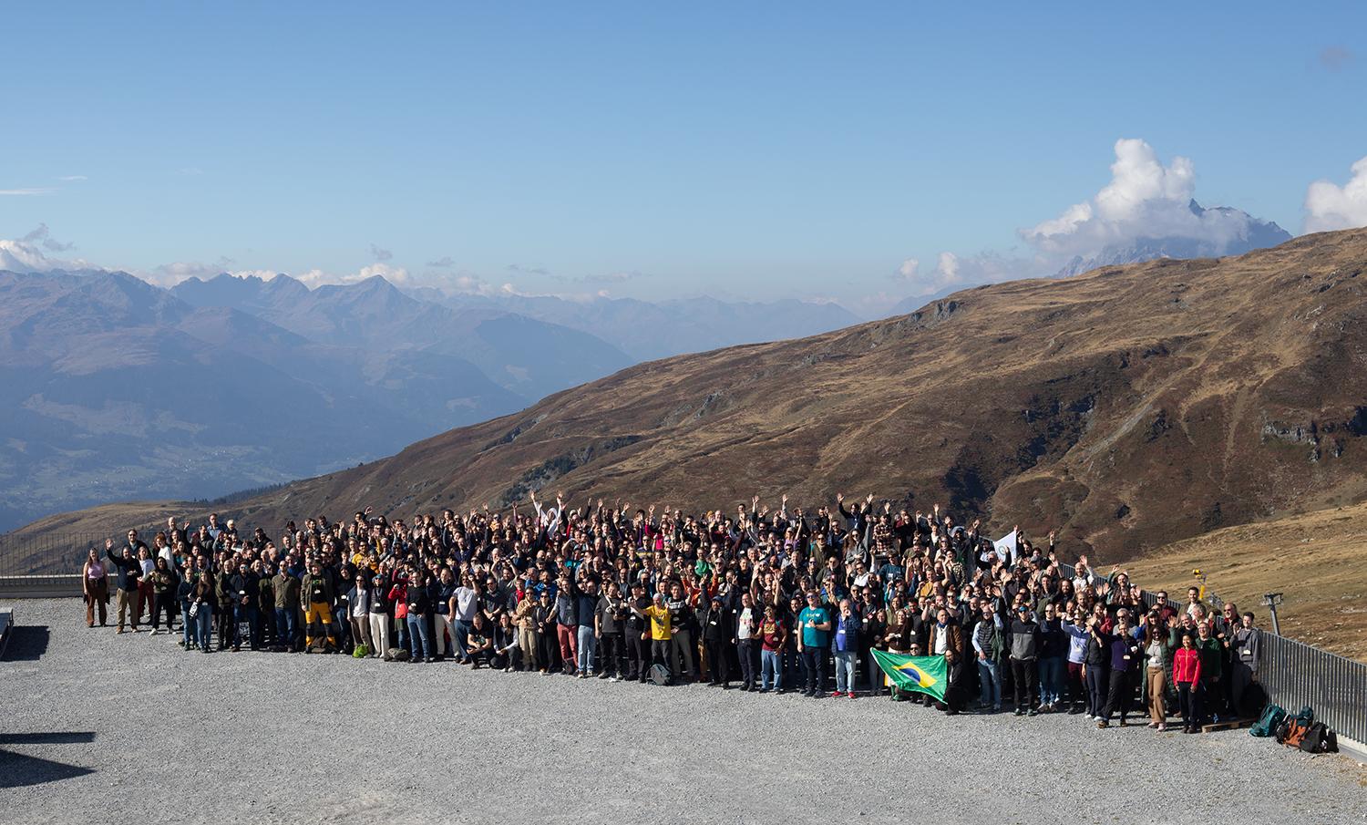 Large group of conference delegates posing on mountain road, holding flags, with vast alpine peaks in background.
